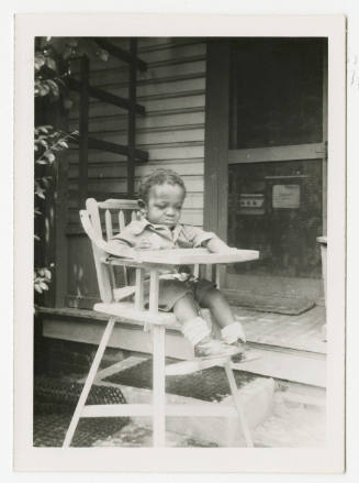 [Young boy sitting in high chair outside]