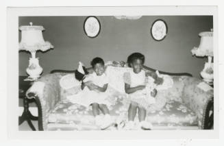 [Two girls with dolls sitting on a sofa]