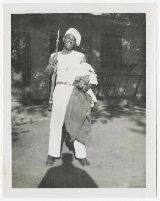 [Man standing in front of house holding a gun, wearing dog tags, Baltimore]