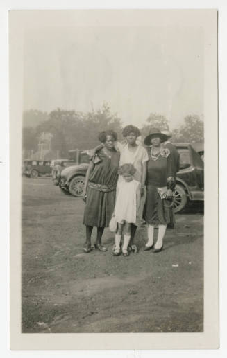 [Three women and a girl standing in front of cars]