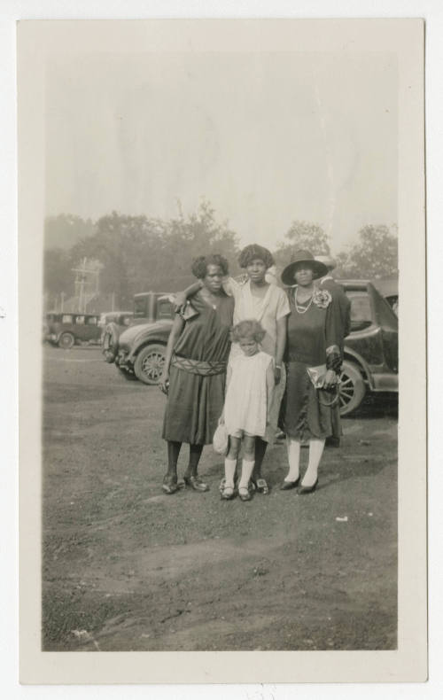 [Three women and a girl standing in front of cars]